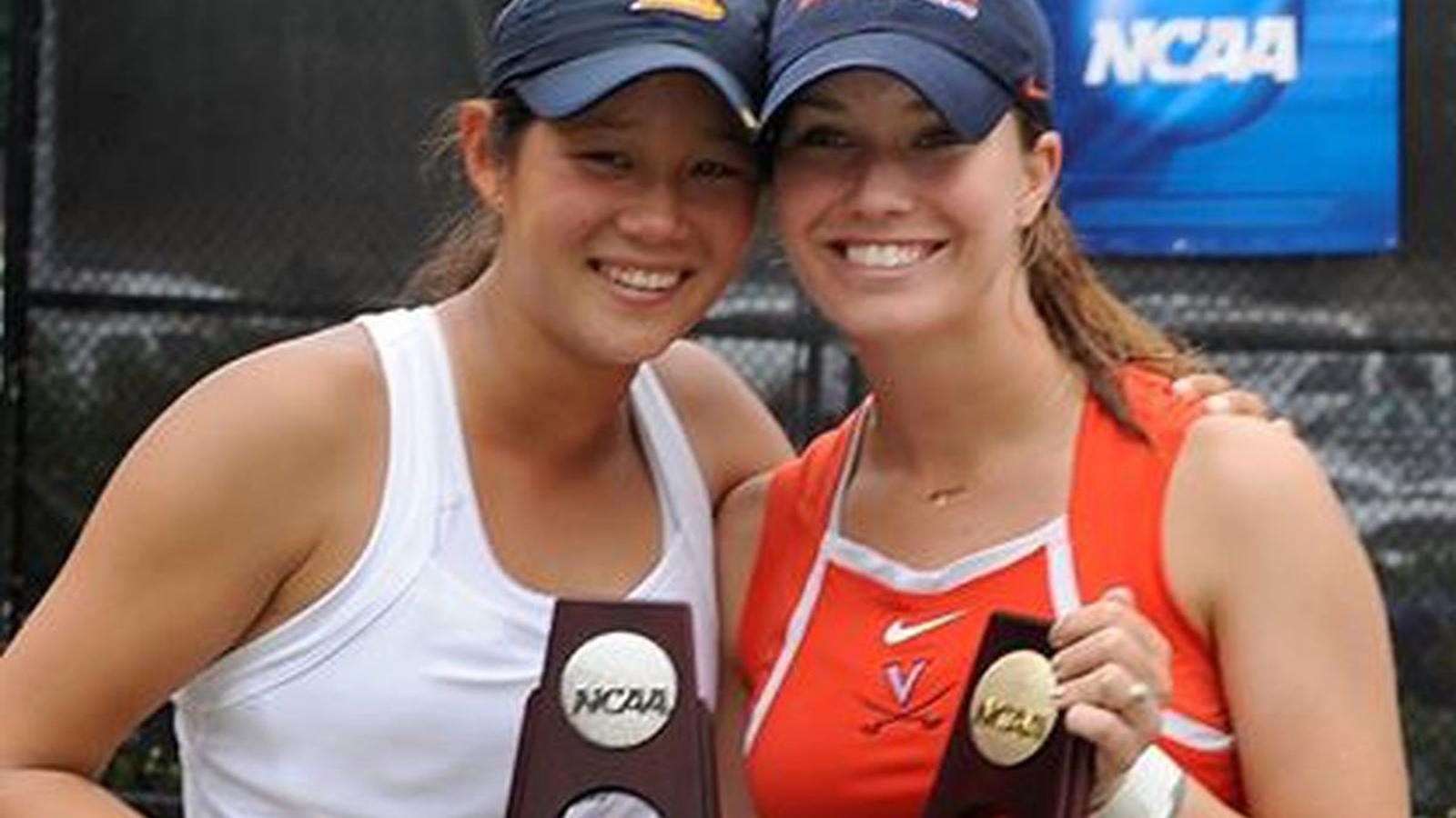 Two smiling female tennis players wearing caps holding NCAA championship trophies together