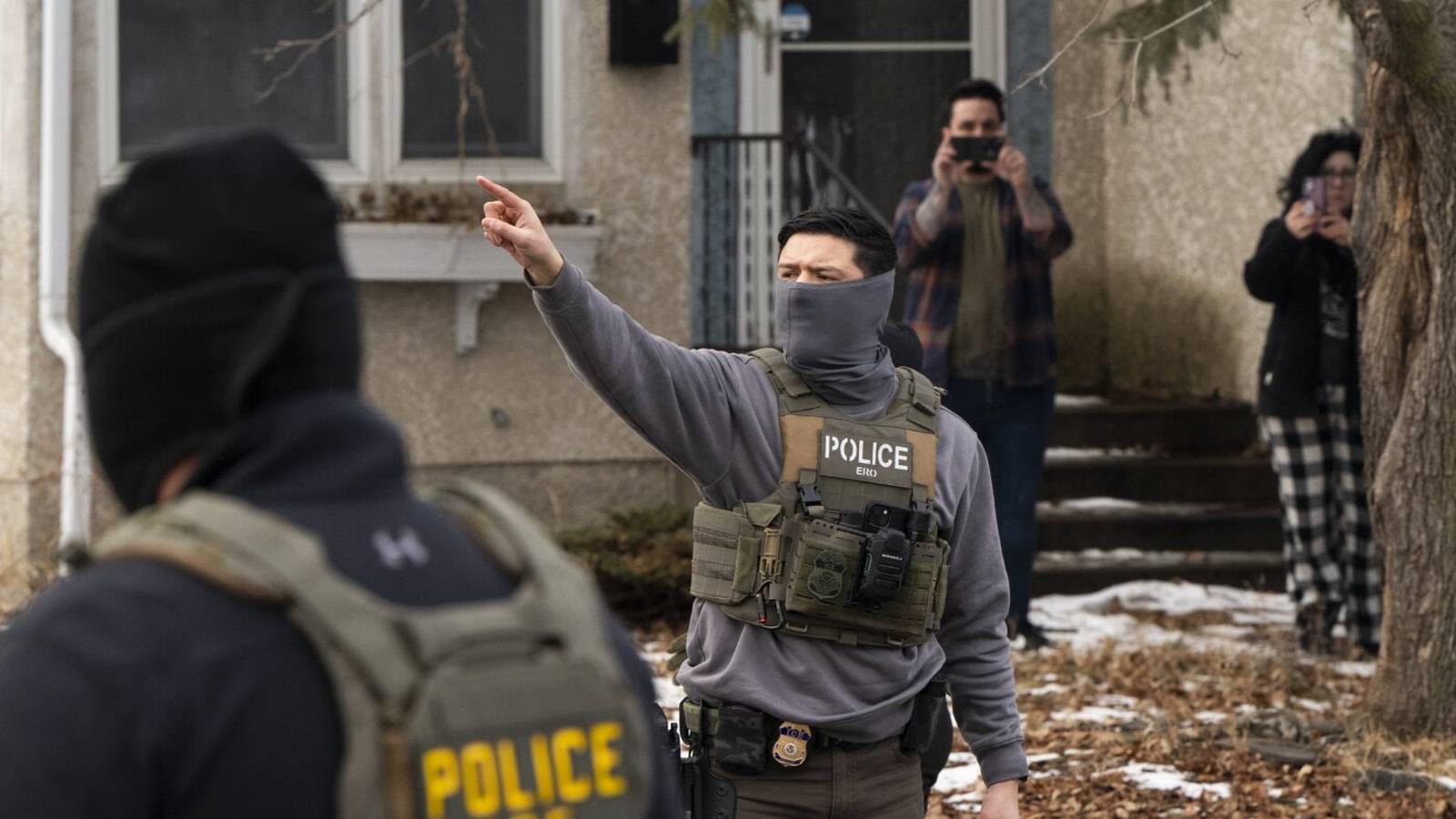 An ICE agent wearing a face covering and tactical vest points toward a house