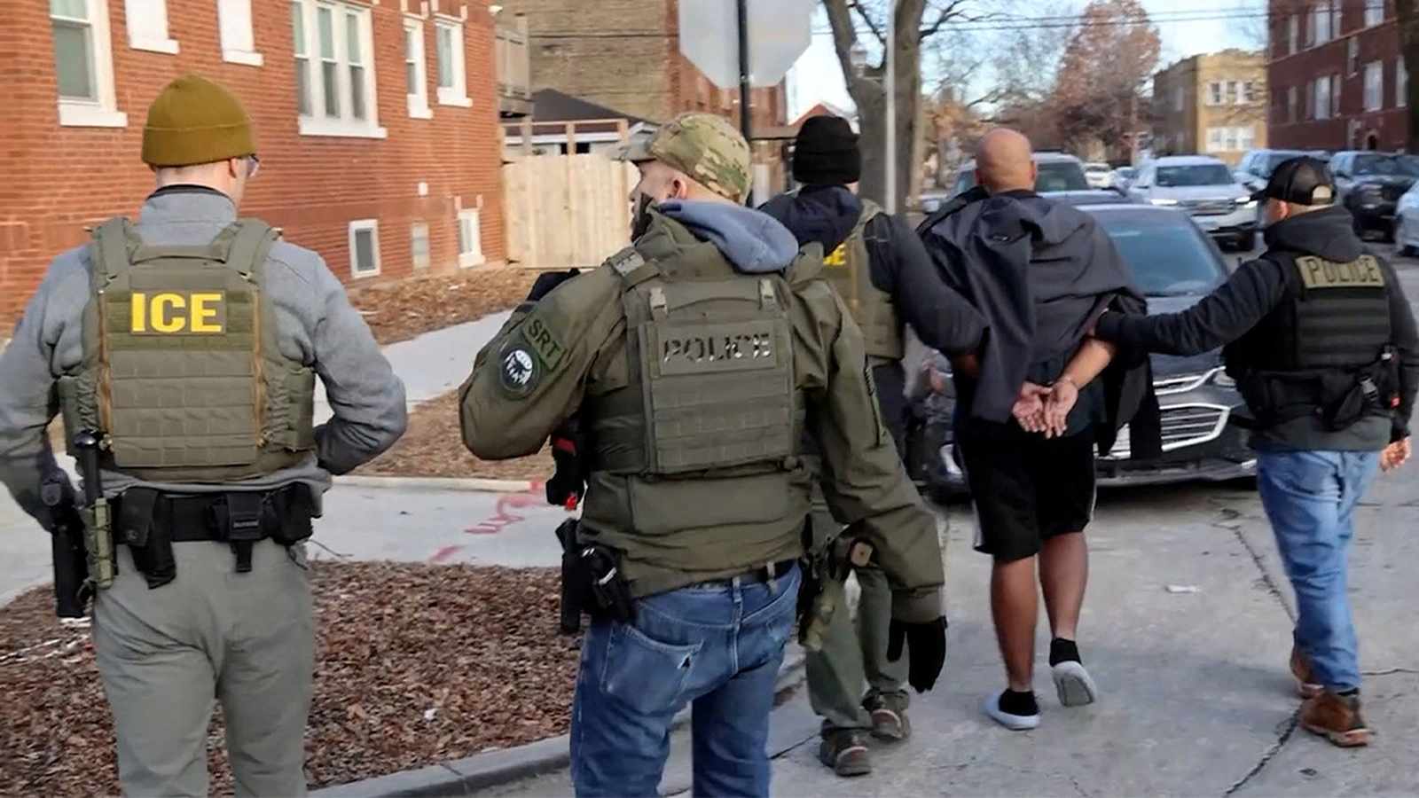 Armed ICE and police officers escorting a handcuffed man down a city street