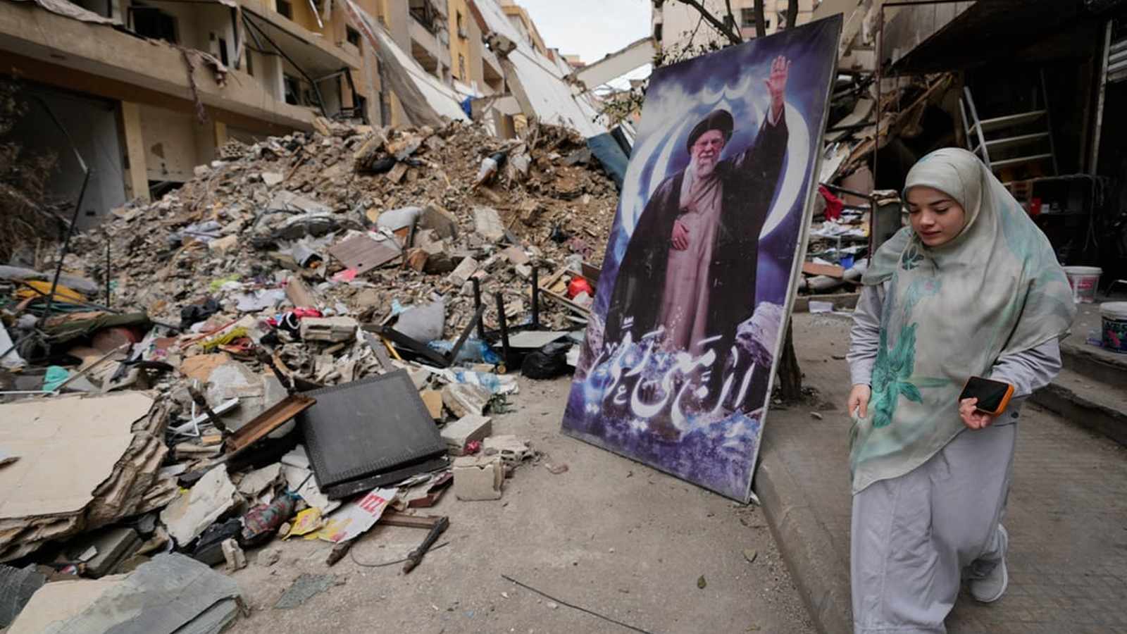 A woman walks past a portrait of Ayatollah Ali Khamenei amidst building rubble