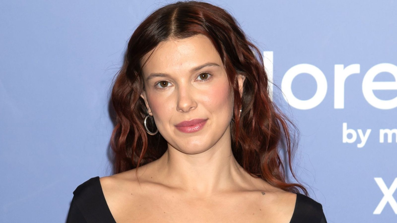 Millie Bobby Brown with long wavy auburn hair posing against a blue background