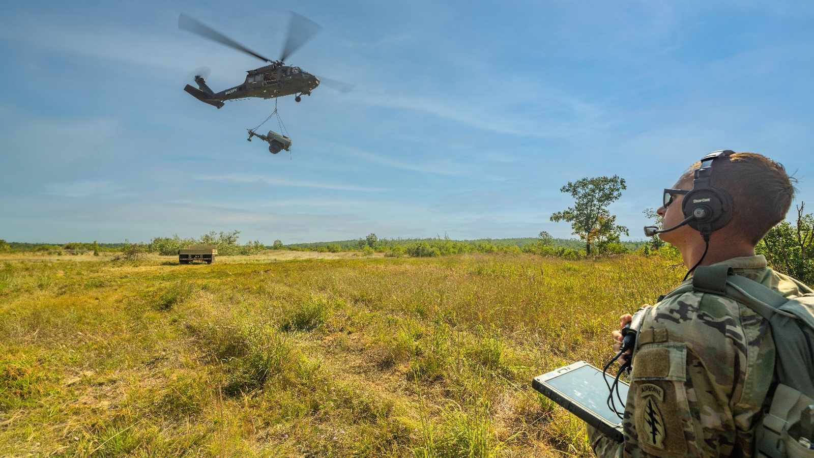 A soldier monitors a military helicopter performing a sling load operation in a field.