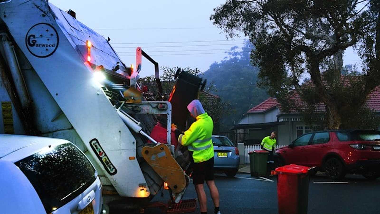 A sanitation worker in high visibility clothing operating a waste collection truck on street