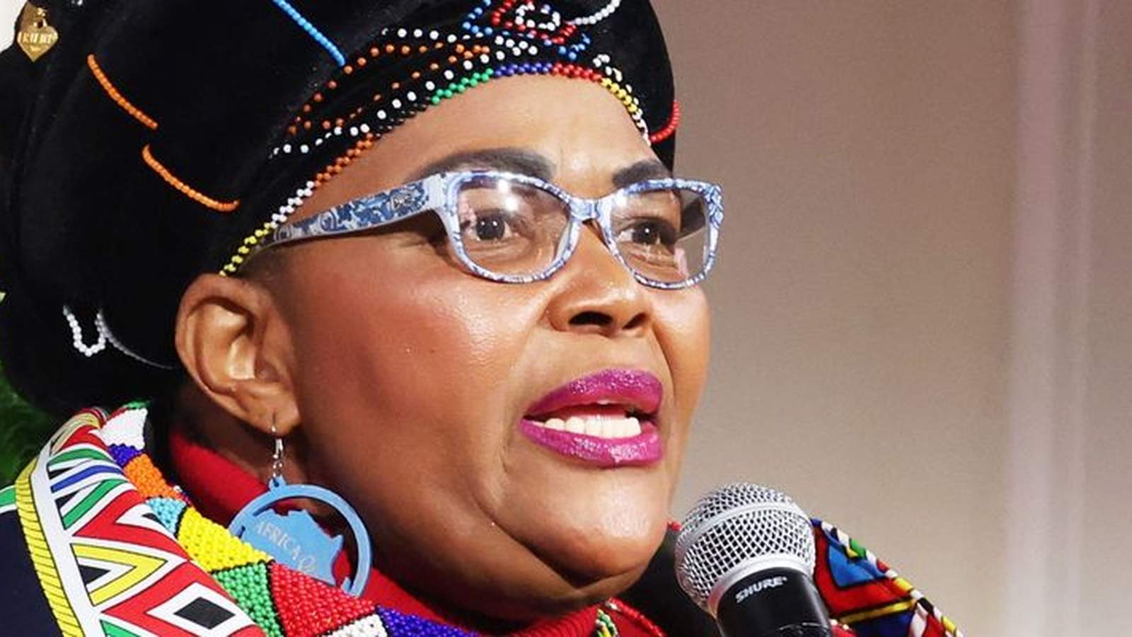 South African Minister Pemmy Majodina wearing traditional attire and glasses speaking into microphon