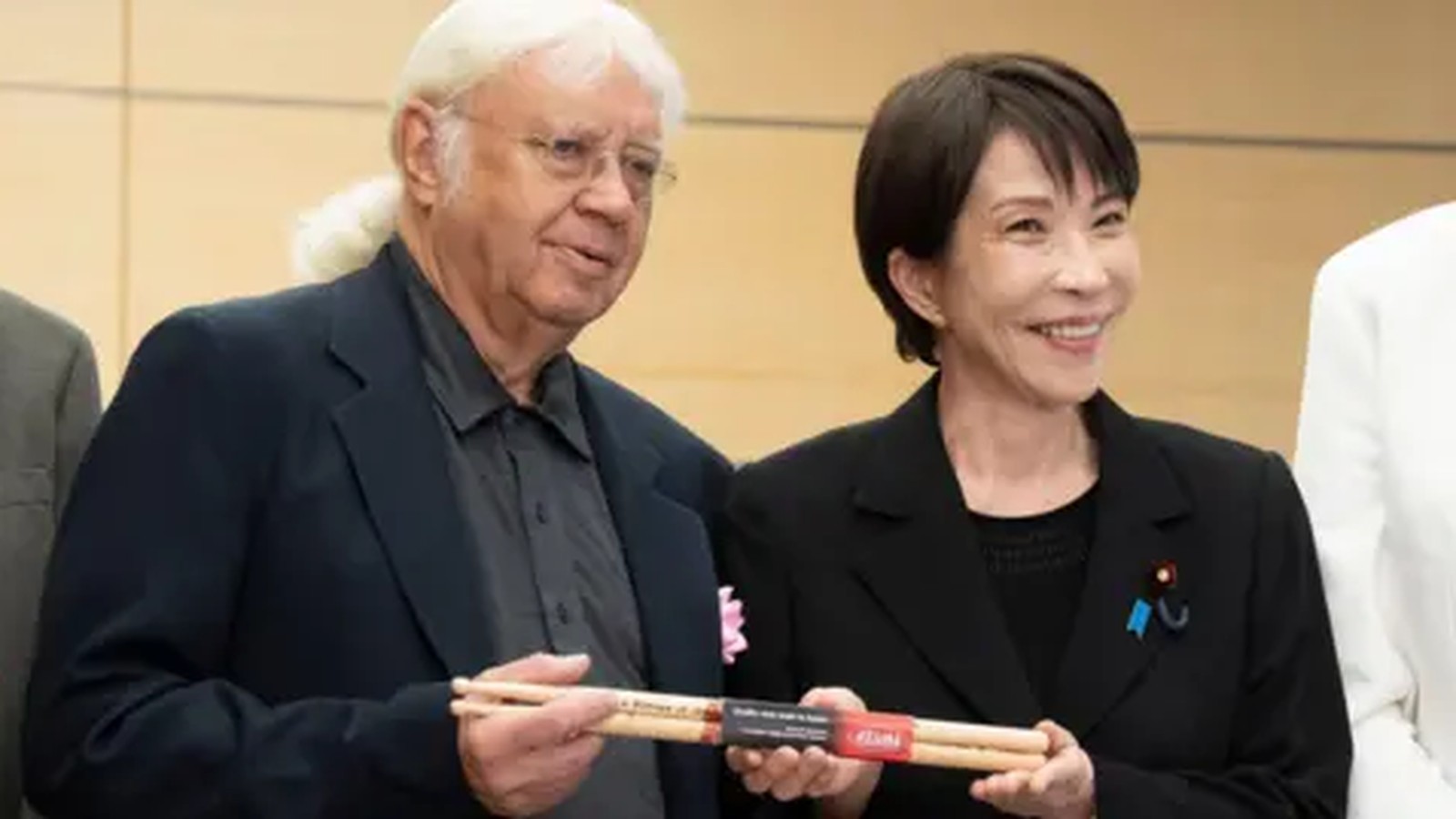 Japanese Prime Minister Sanae Takaichi smiling while holding drumsticks with Deep Purple drummer Ian