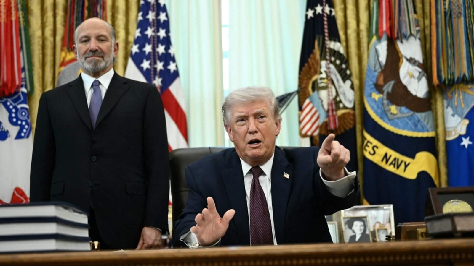 Donald Trump sits at a desk pointing while an official stands behind him
