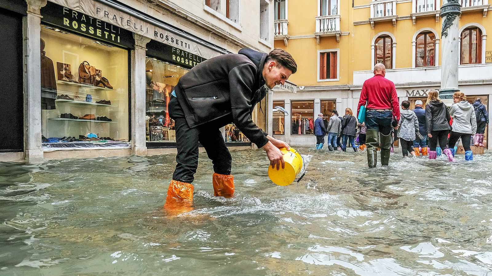 A man in orange boots scoops water from a flooded street in Venice