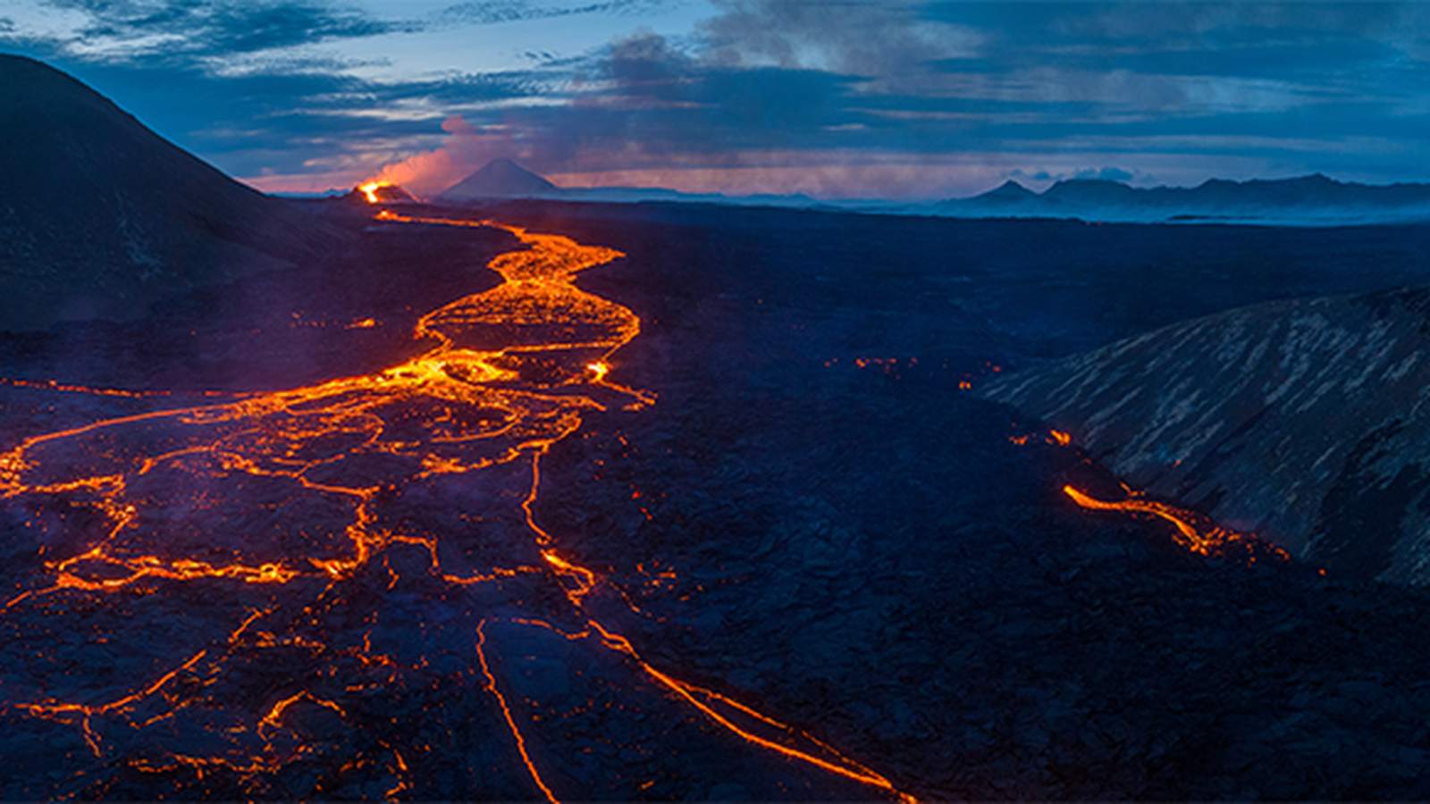 A panoramic view of glowing orange lava flowing through a dark volcanic landscape at twilight