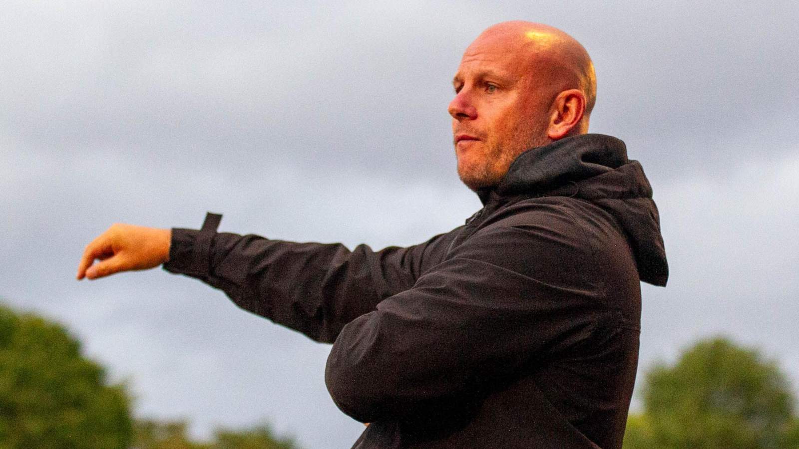 Football manager Adam Murray standing on the sidelines wearing a black jacket