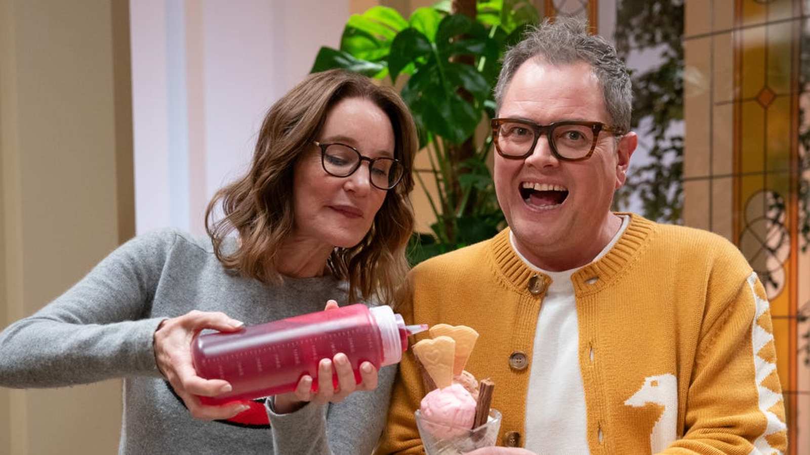 Alan Carr and Tina Fey smile while sharing an ice cream sundae with syrup