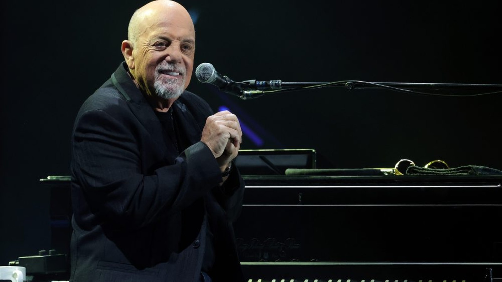Billy Joel smiling while performing at a grand piano on a dark stage