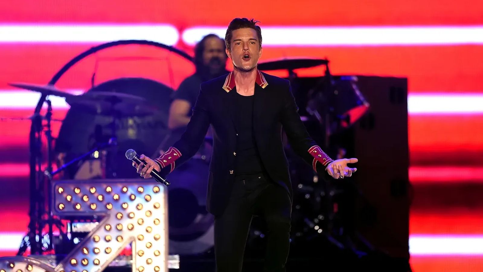 Brandon Flowers singing into a microphone on stage with a drum kit behind him