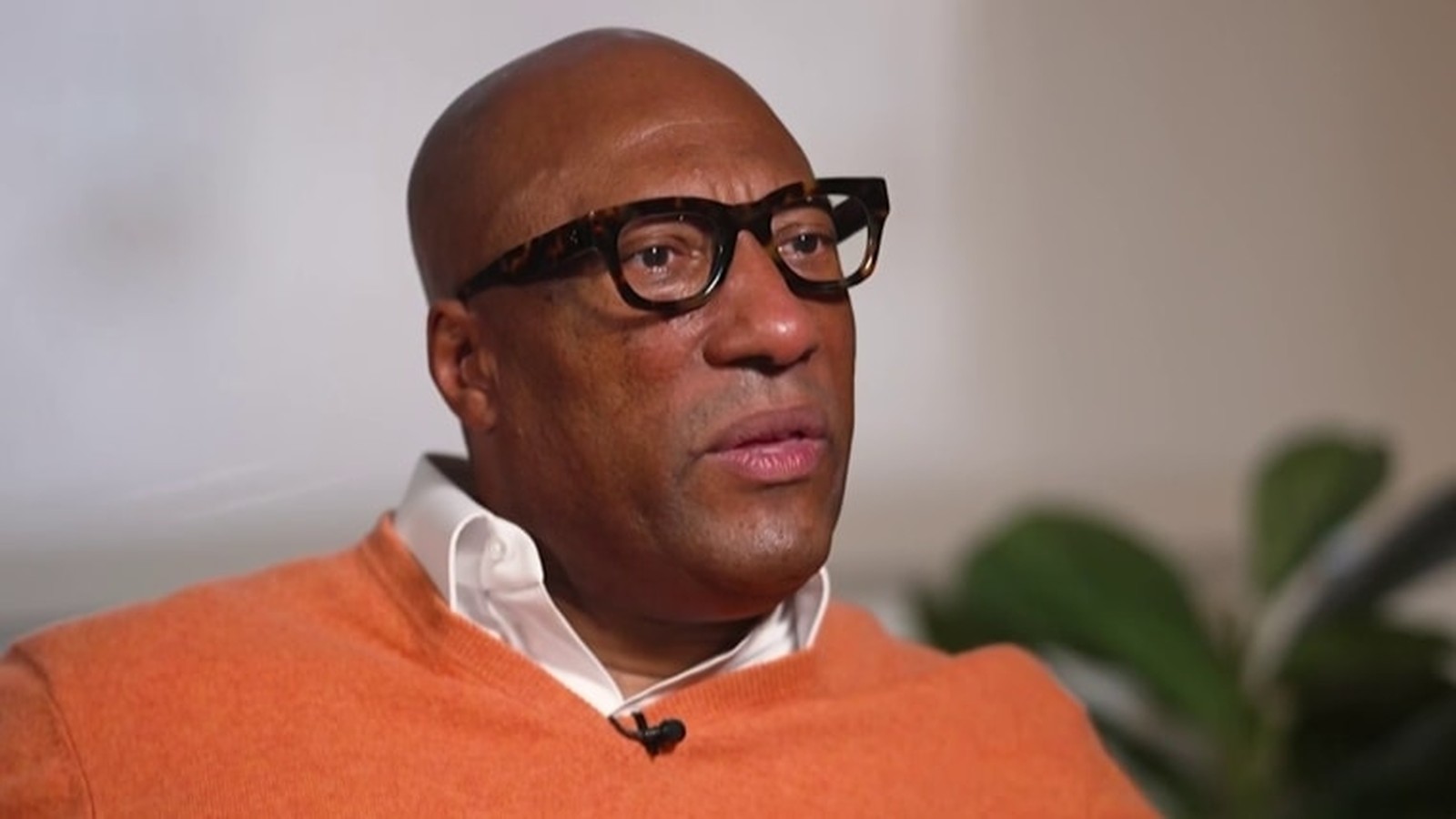 Byron Allen wearing thick-rimmed glasses and an orange sweater during a professional interview
