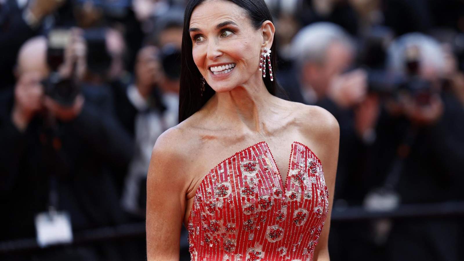 Demi Moore smiling on a red carpet wearing a red beaded gown