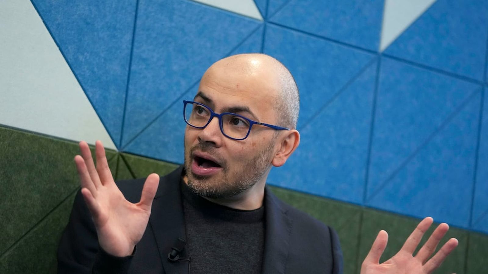 Demis Hassabis wearing blue glasses gesturing while speaking against a geometric blue background