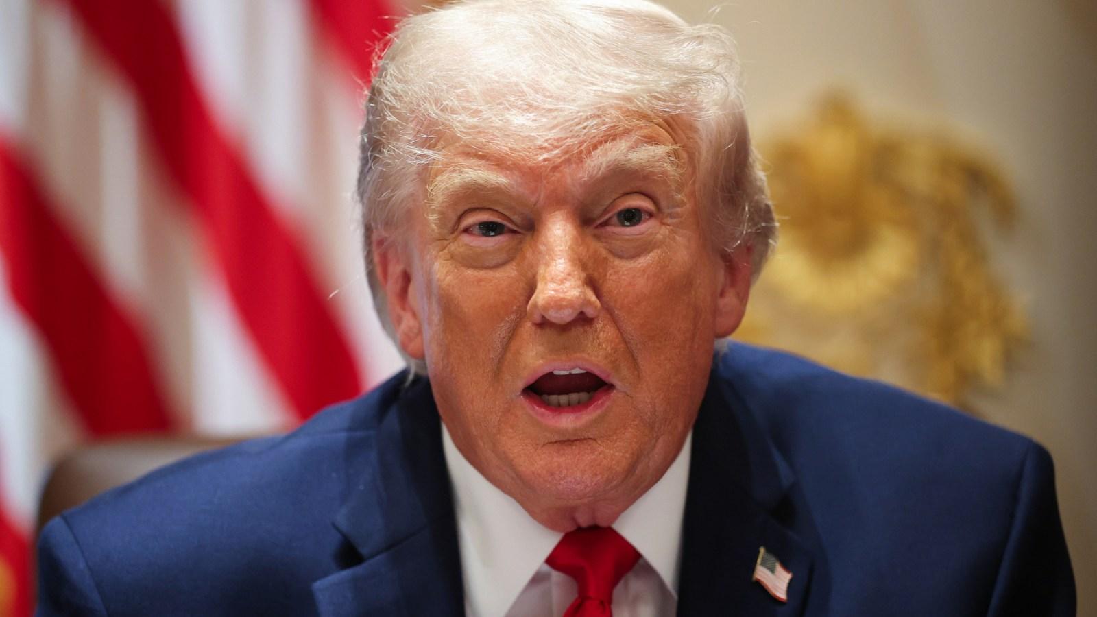 Donald Trump speaking with a serious expression in front of an American flag