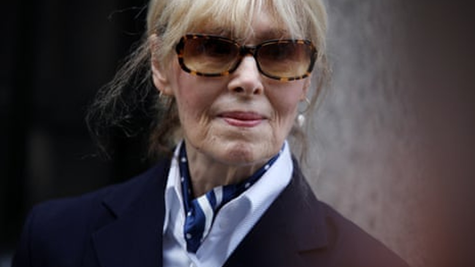 E. Jean Carroll wearing sunglasses and a blue blazer with a neck scarf