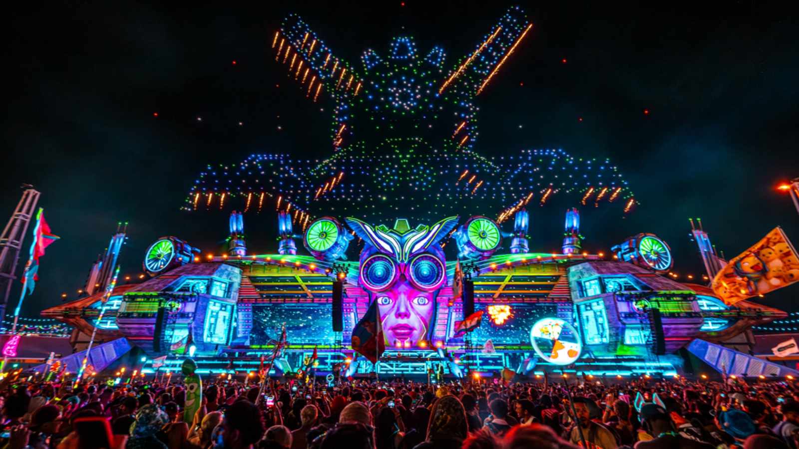 Massive illuminated stage with owl face design and drone show at night festival