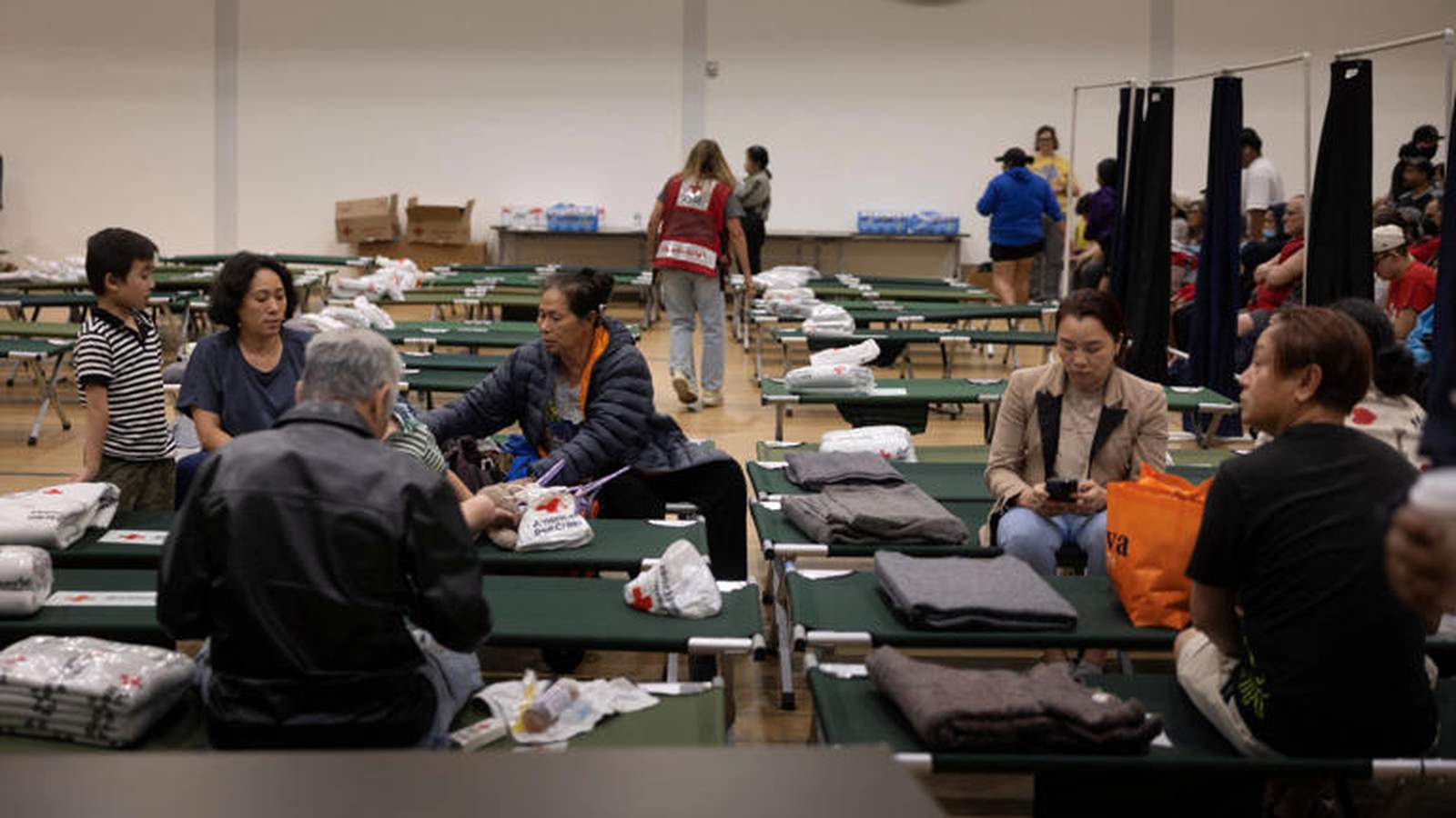 People sitting on cots inside an emergency shelter with Red Cross supplies visible