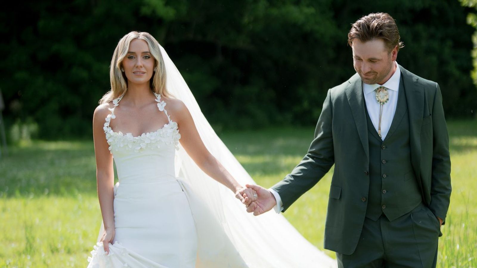 Lainey Wilson in a white wedding dress holding hands with Devlin Hodges outdoors