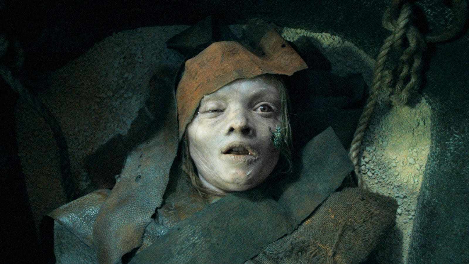 A pale, mummified figure with a beetle on its cheek in a dark tomb