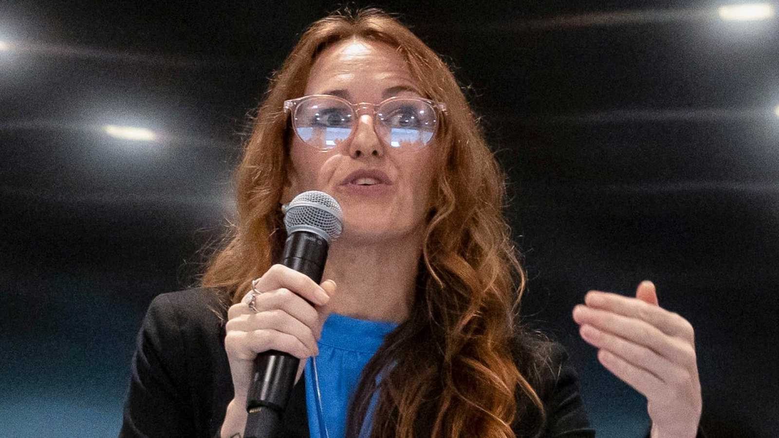 Maureen Galindo with long hair and glasses speaking into a microphone at event