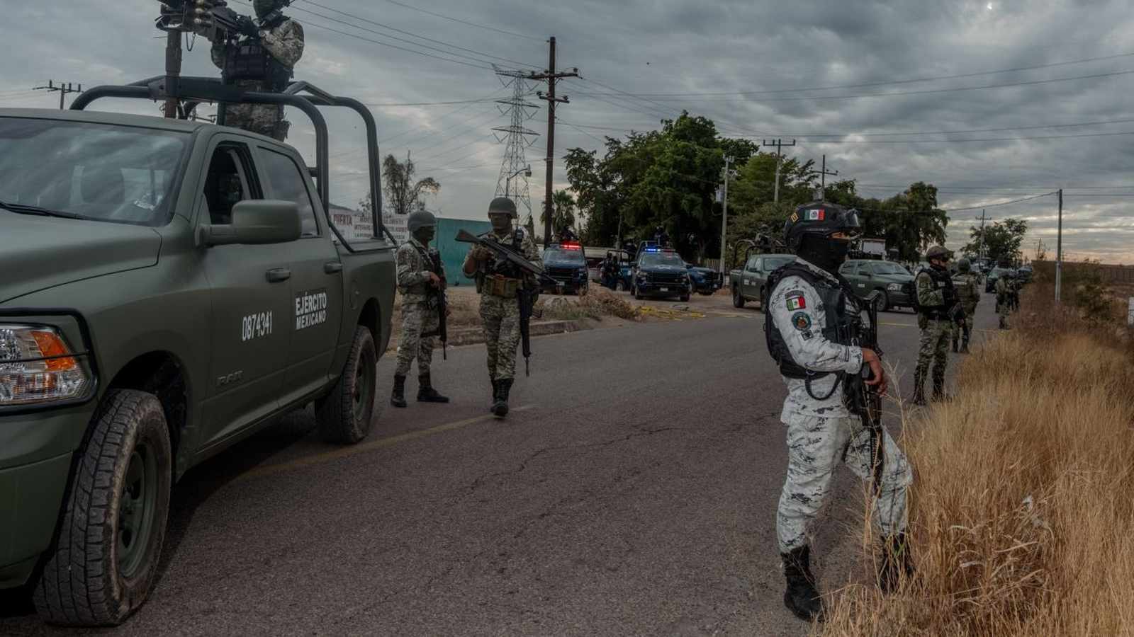 Mexican military personnel in tactical gear stand guard near armed vehicles on a road under a cloudy