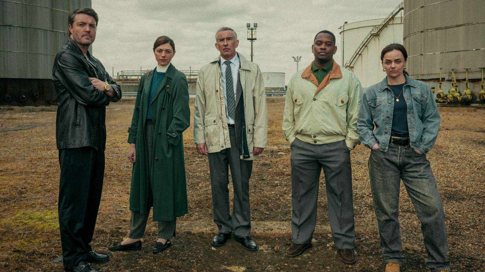 Five actors standing in a line outdoors in front of industrial storage tanks