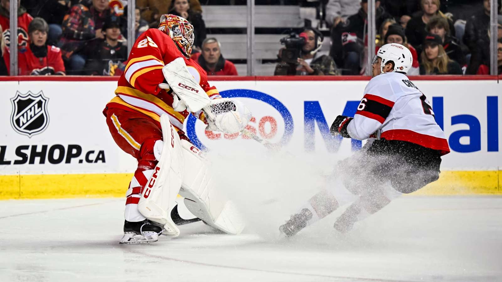 A hockey goalie in red and yellow makes a save as a player in a white jersey skates by, kicking up s