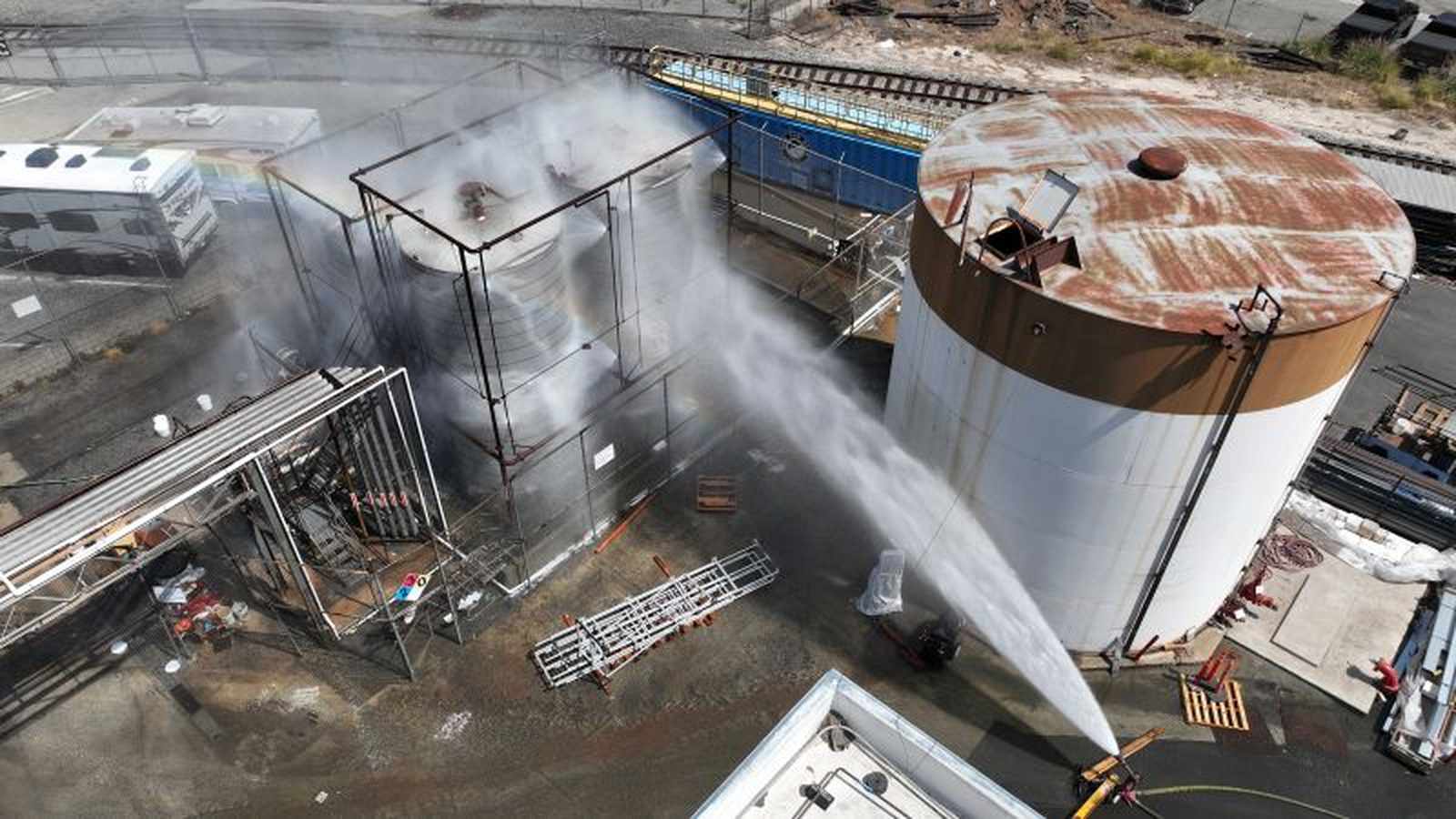 Aerial view of firefighters spraying water on a large industrial chemical tank