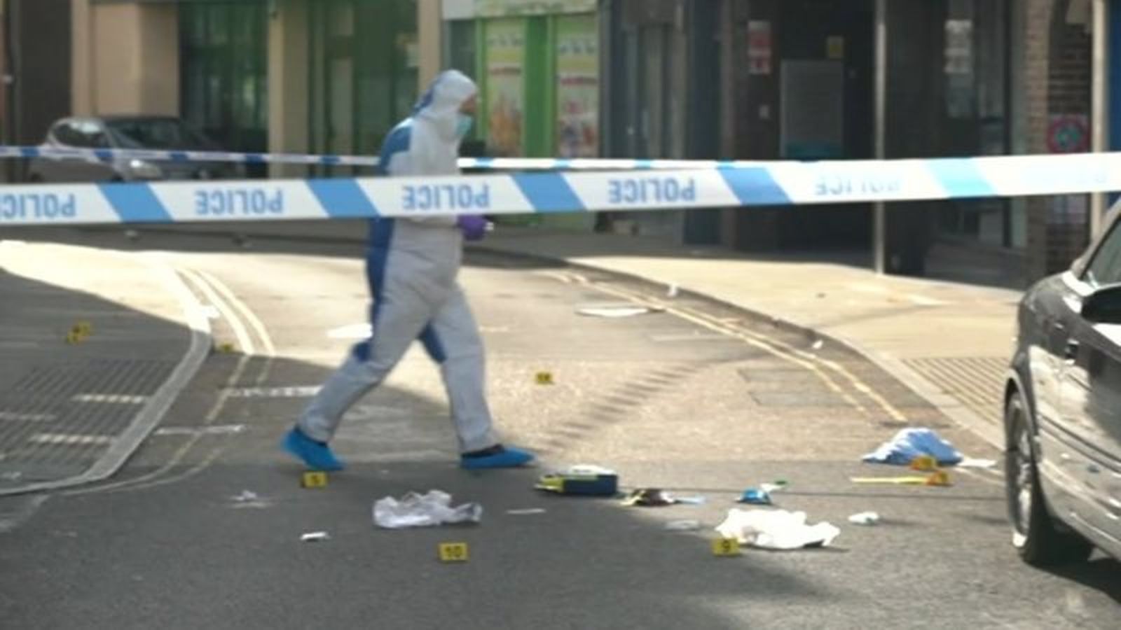 A person in a forensic suit walks past police tape at a street crime scene with evidence markers
