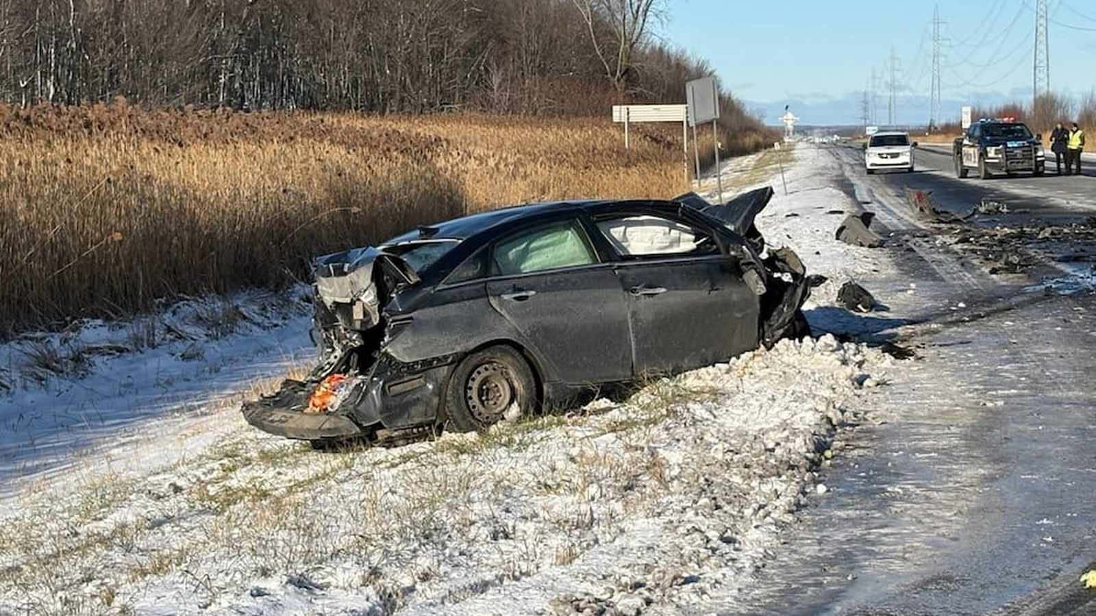 A heavily damaged black car sits in a snowy ditch beside a highway