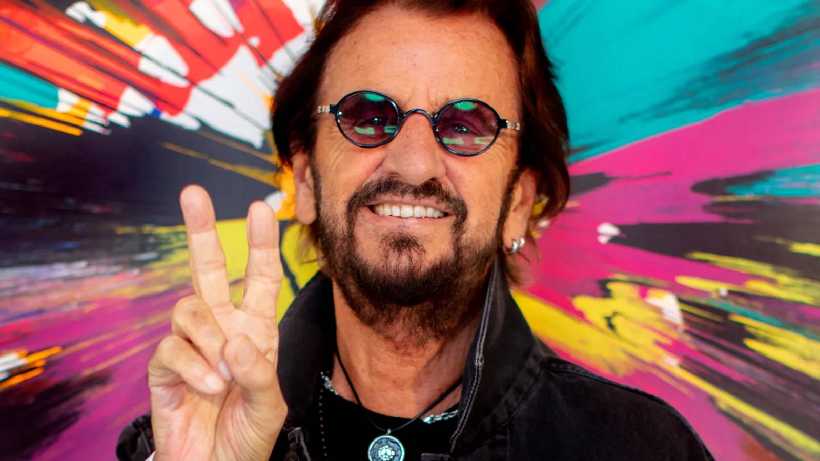 Ringo Starr smiling and making a peace sign against a colorful abstract background