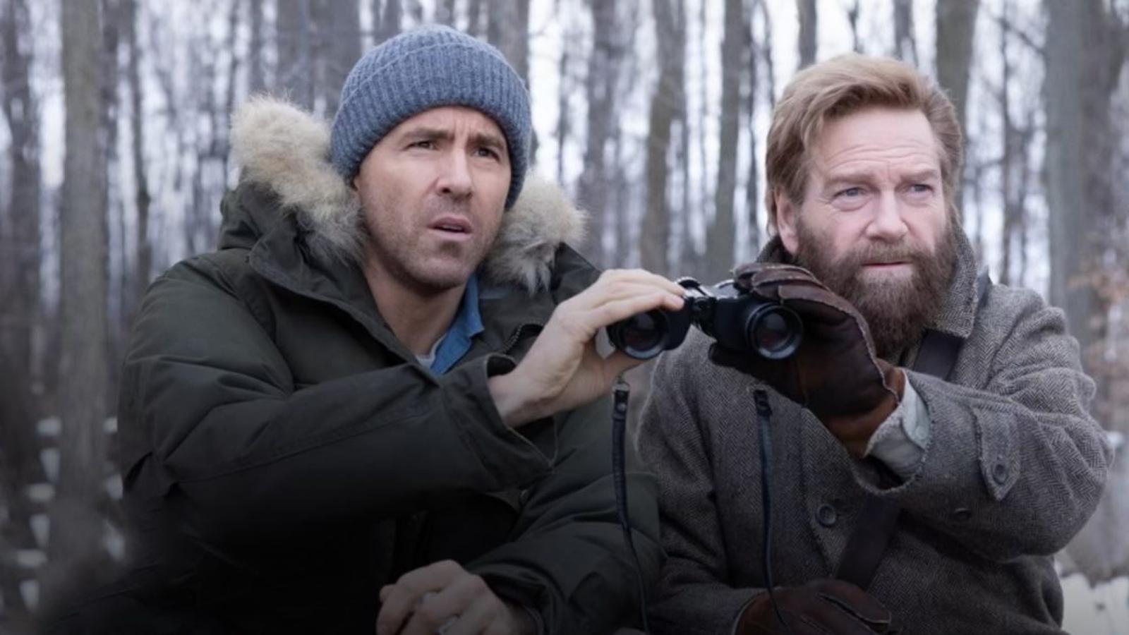 Ryan Reynolds wearing a winter parka and beanie looking through binoculars in woods