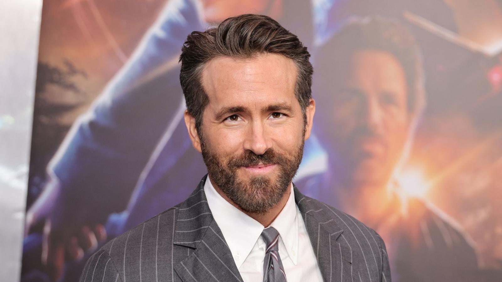 Ryan Reynolds wearing a grey pinstripe suit jacket and tie smiling at camera