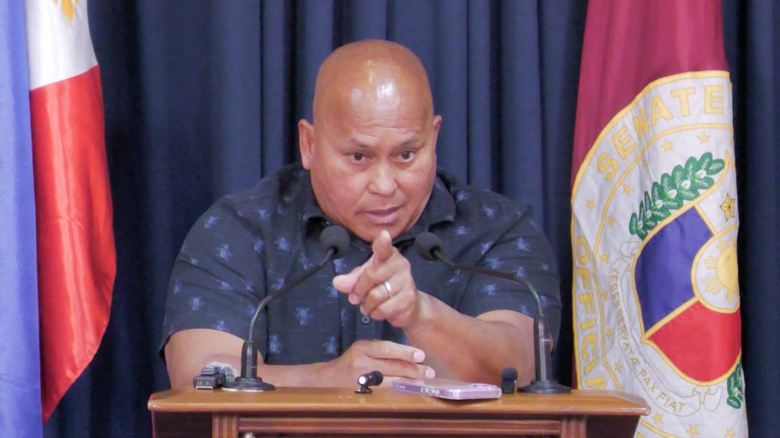 Philippine Senator Ronald Dela Rosa points forward while speaking at a press conference with flags b
