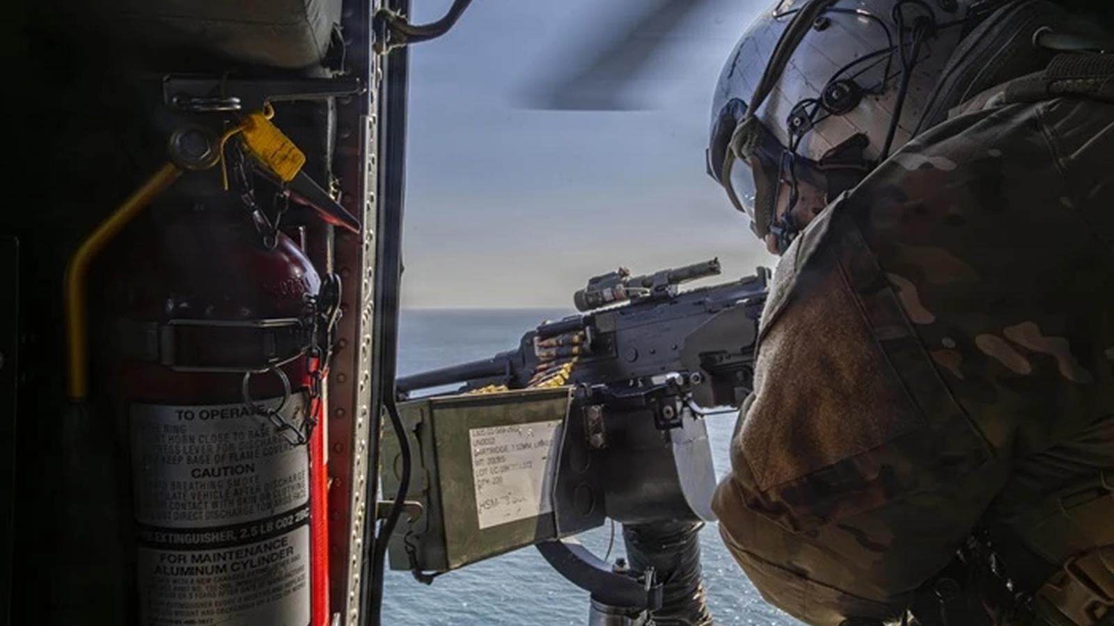 A US Navy helicopter crew member operates a mounted machine gun over the ocean.