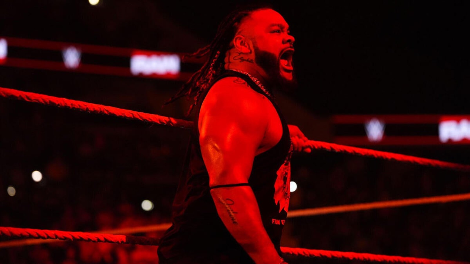 A male wrestler with braided hair and tattoos shouts in a WWE wrestling ring under red lighting