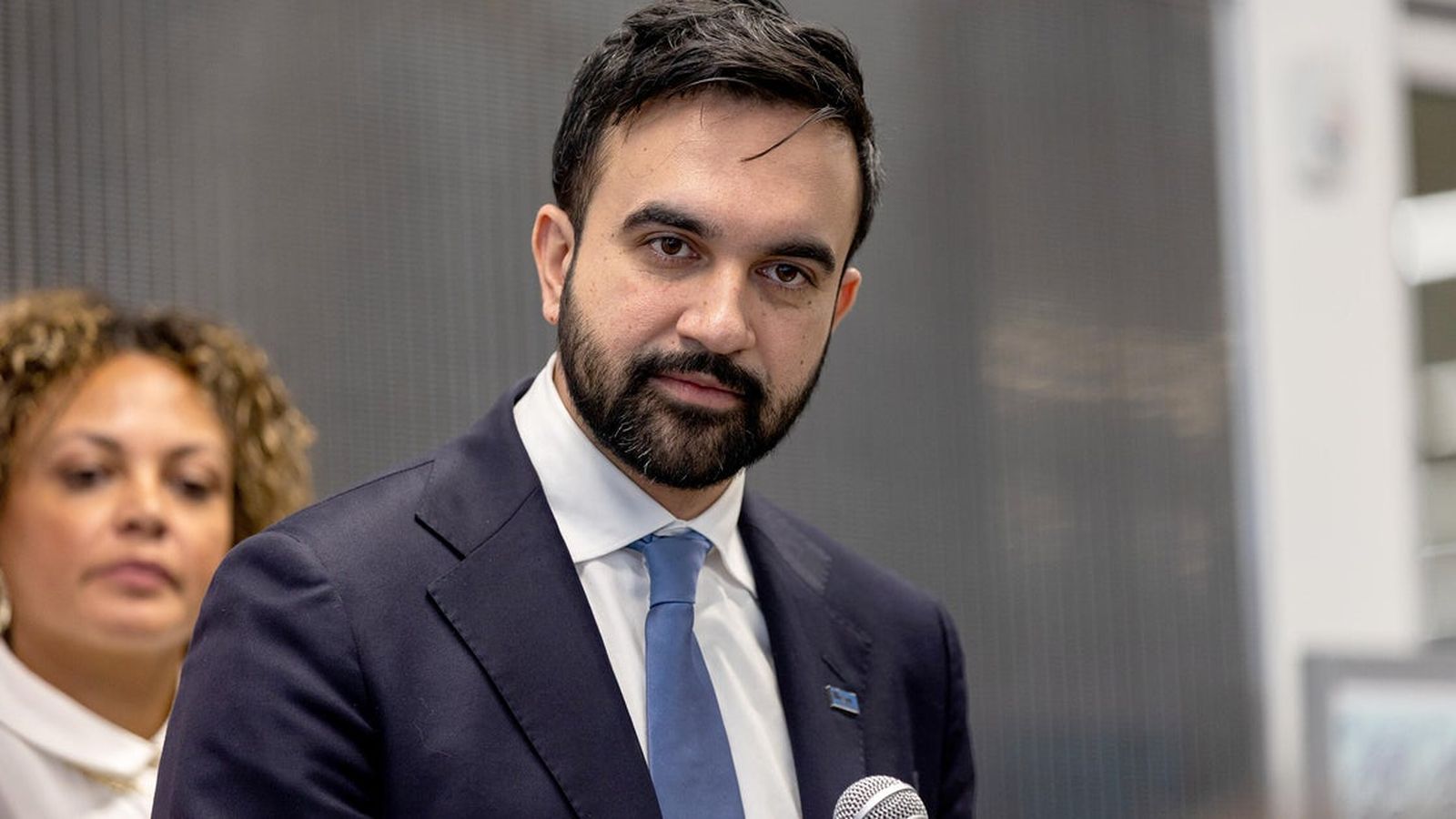 Zohran Mamdani wearing a suit and blue tie speaking at a press conference