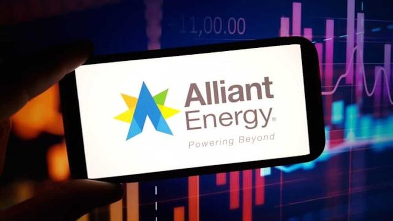 A hand holding a smartphone displaying the Alliant Energy logo against a stock chart