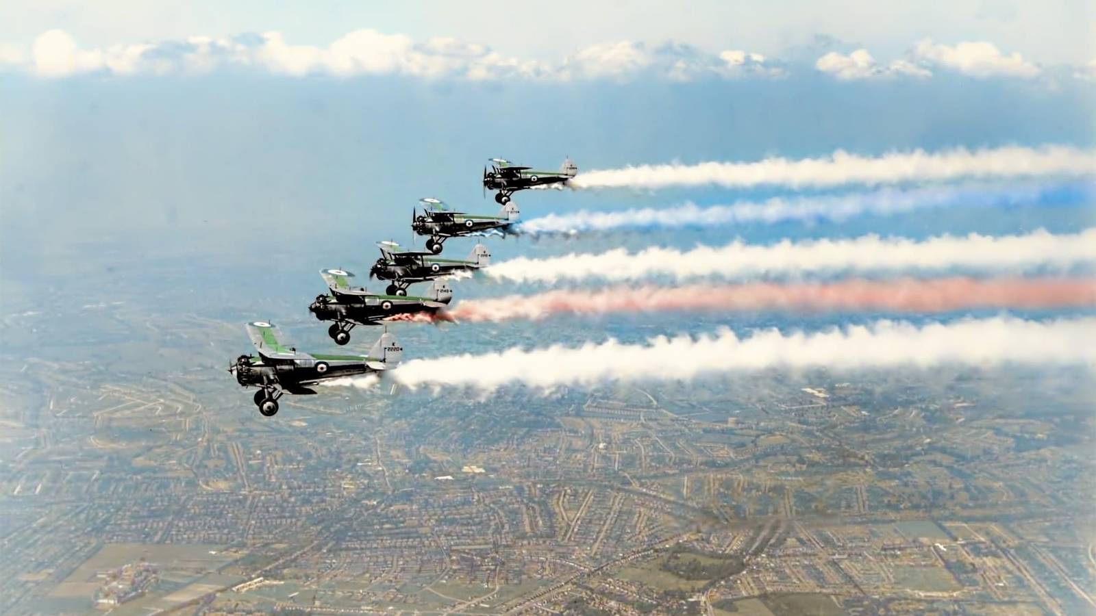 Five vintage biplanes flying in formation leaving red white and blue smoke trails