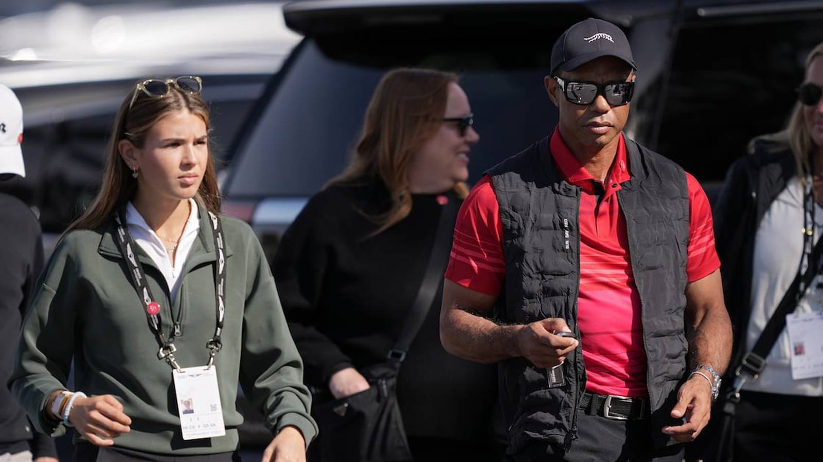 Tiger Woods wearing sunglasses and a black vest walking with a young woman