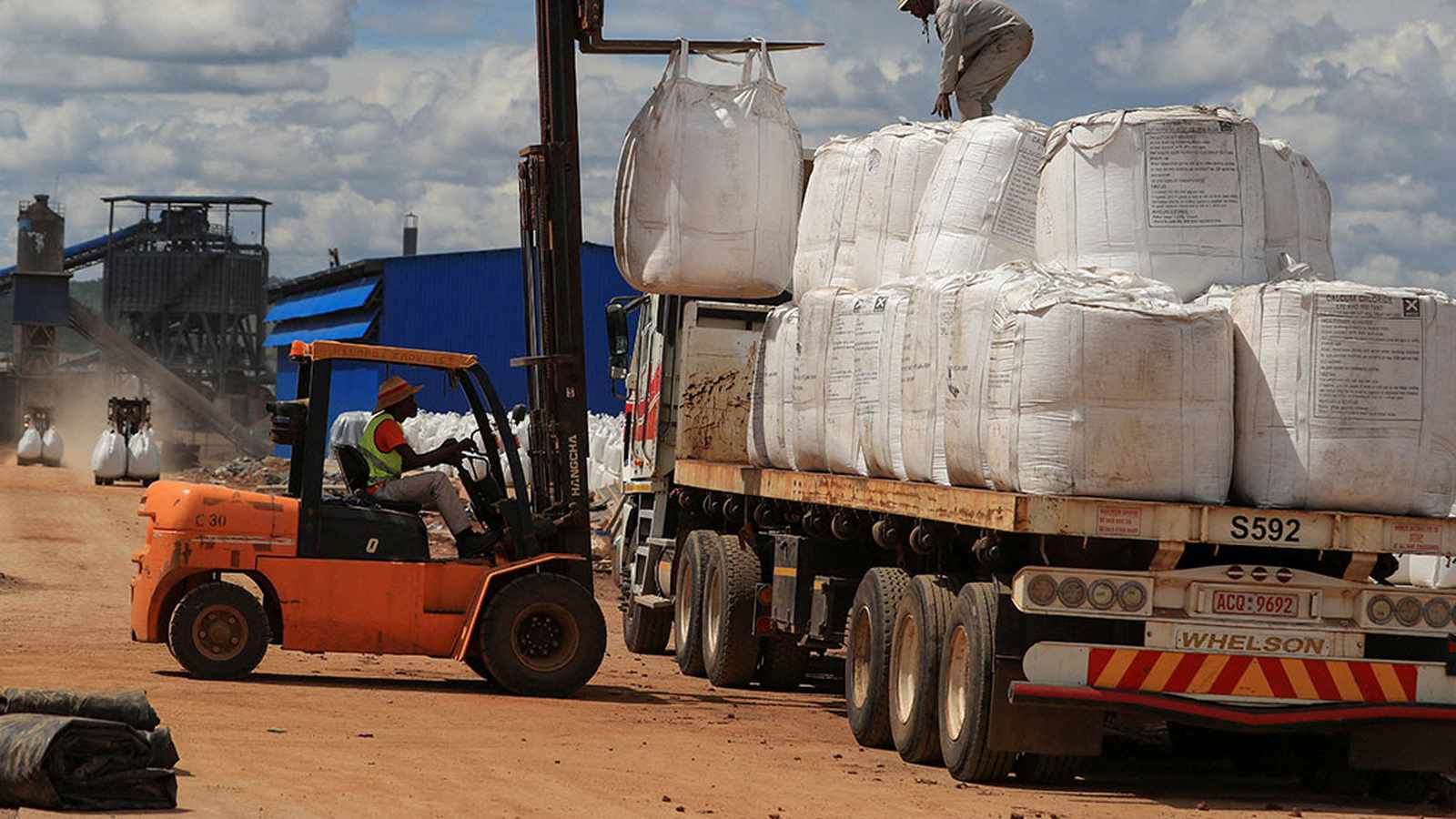 A forklift operator loading large white mineral bags onto a flatbed truck