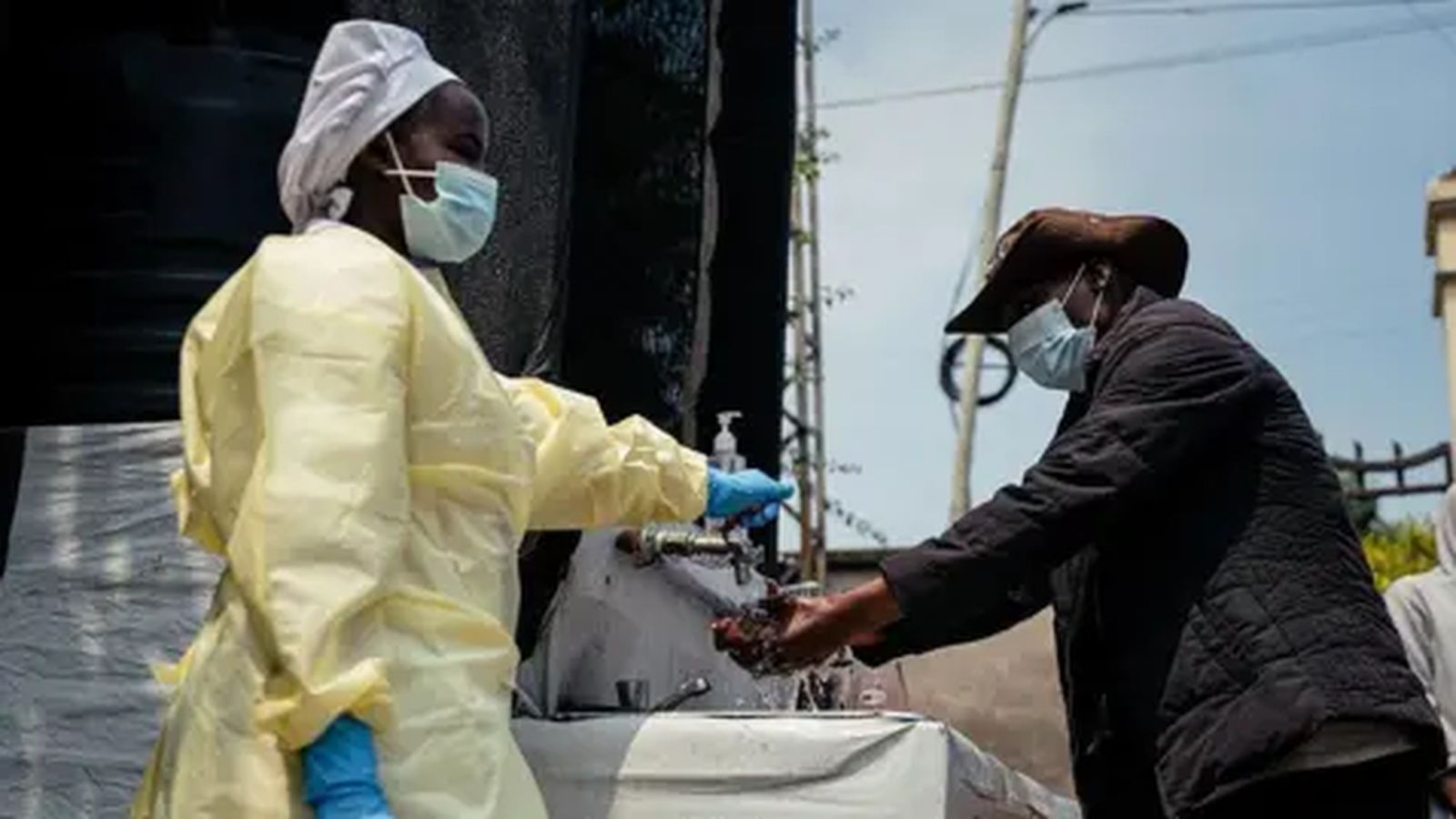 A health worker in protective gear supervises a man washing his hands outdoors