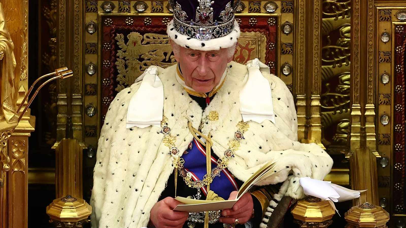 King Charles III wearing a crown and ermine robe reads from a document