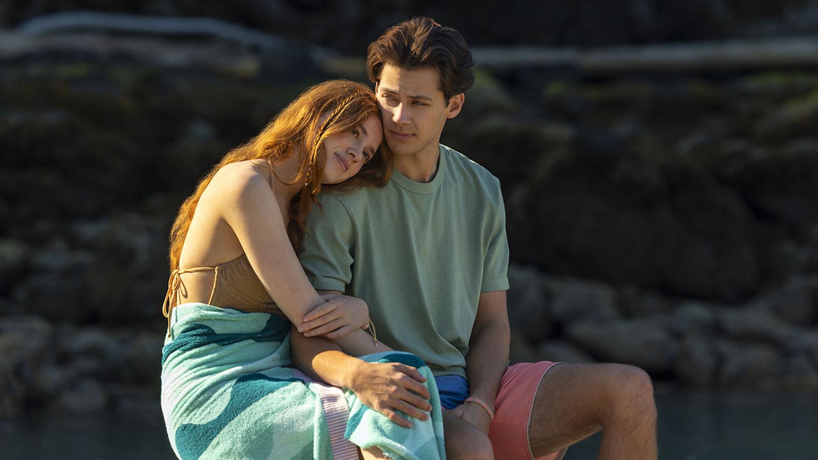 A young woman with long red hair rests her head on a mans shoulder, both sitting outdoors