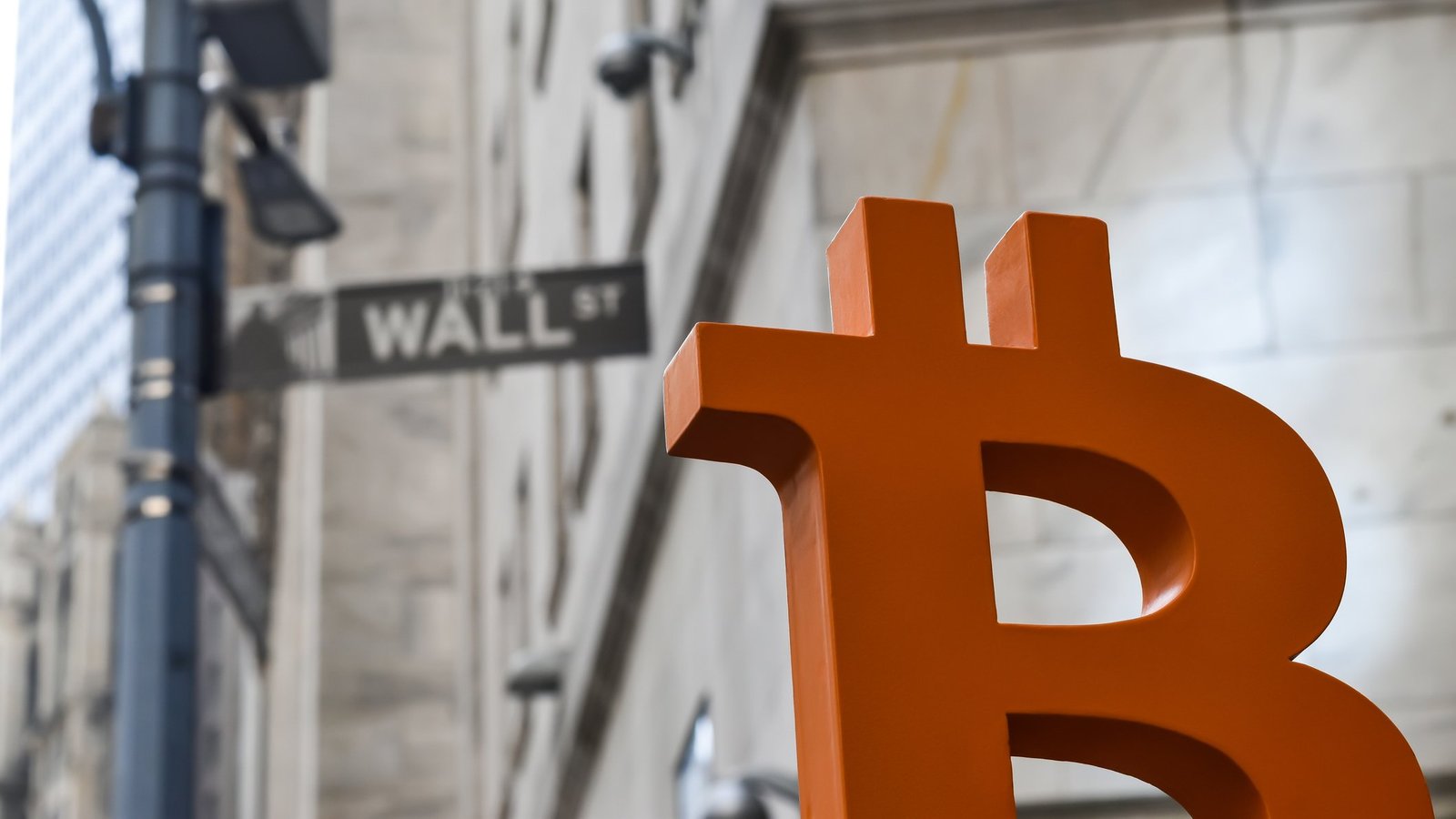 A large orange Bitcoin symbol sculpture positioned in front of a Wall Street sign