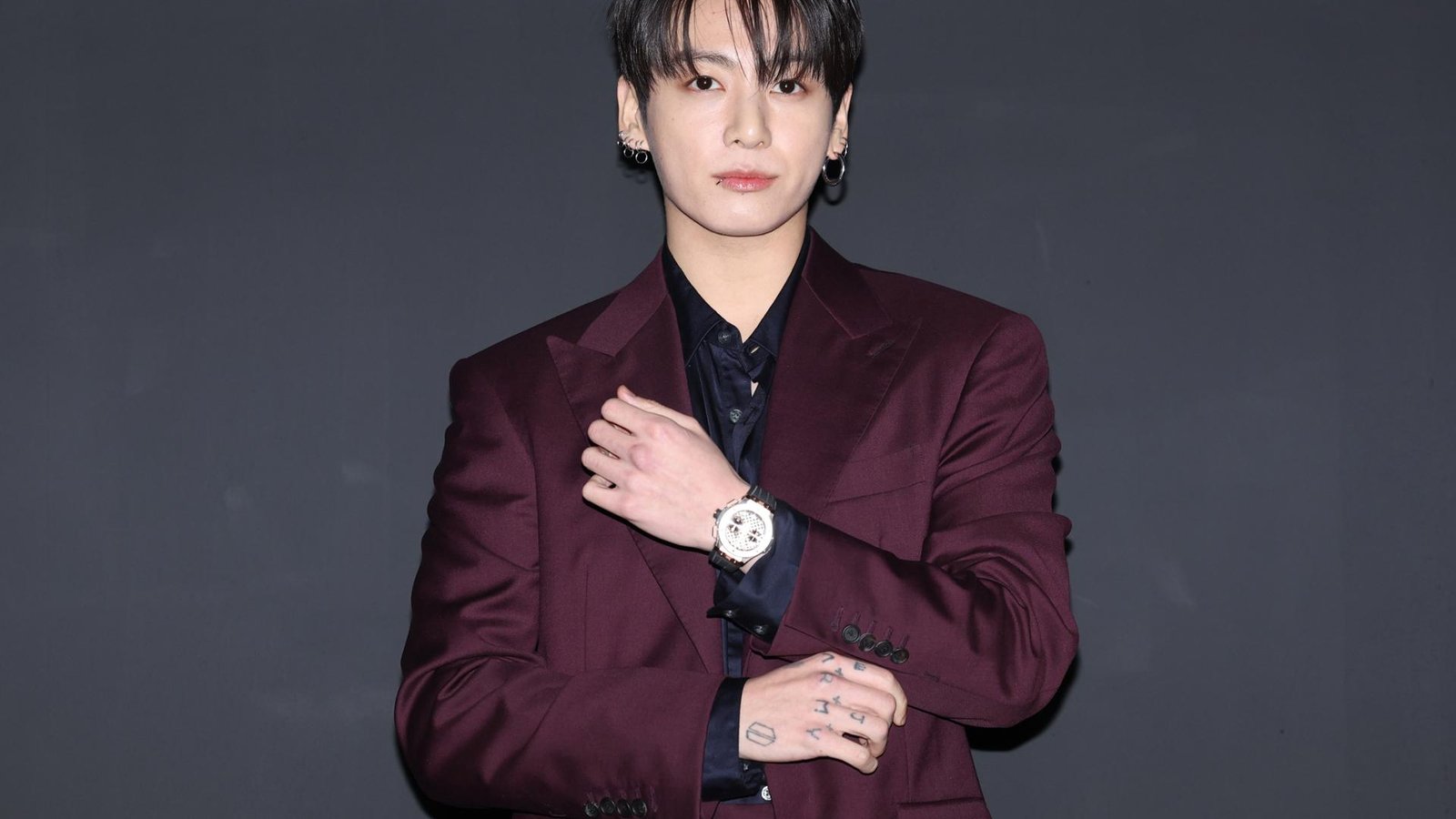 BTS member Jungkook wearing a dark burgundy suit against a plain grey background
