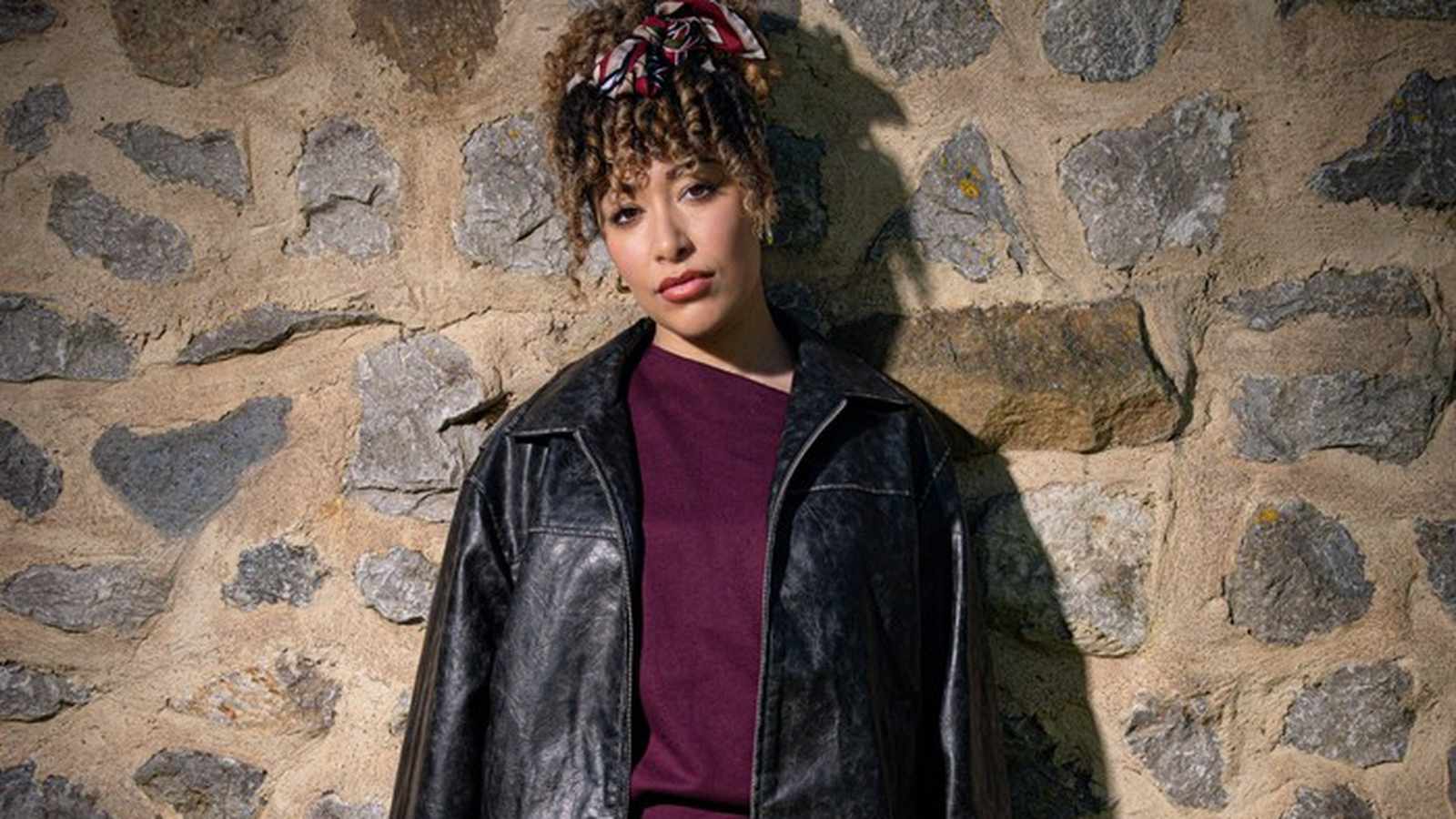 Actress Casey Al-Shaqsy posing against a rustic stone wall for her Emmerdale role