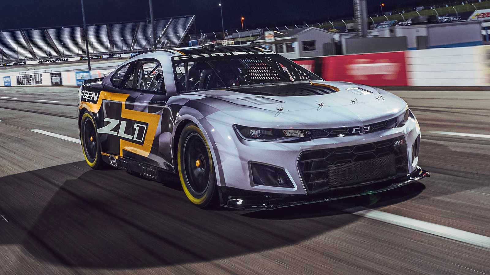 A silver Chevrolet Camaro ZL1 race car speeding on a NASCAR track at night